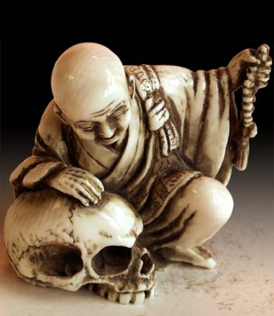Netsuke ( 根付 ) en ivoire : le personnage entièrement rasé s'appuie d'une main sur un crâne sans machoire inférieure et de l'autre main un objet représentant des perles sur un lacet et servant peut-être à compter.  JPEG - 23 ko